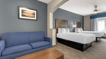 A hotel room with a blue sofa, a wooden coffee table, two beds with white linens, a framed picture on the wall, and a ceiling fan.