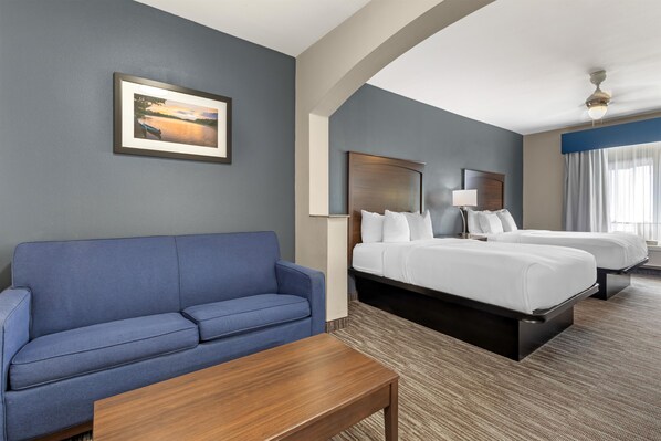 A hotel room with a blue sofa, a wooden coffee table, two beds with white linens, a framed picture on the wall, and a ceiling fan.