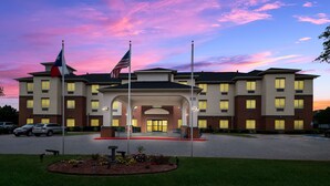 A multi-story hotel with a central entrance, flagpoles, and a landscaped area with a bench and flowers. - Best Western Plus Lake Dallas Inn & Suites (Lake Dallas)