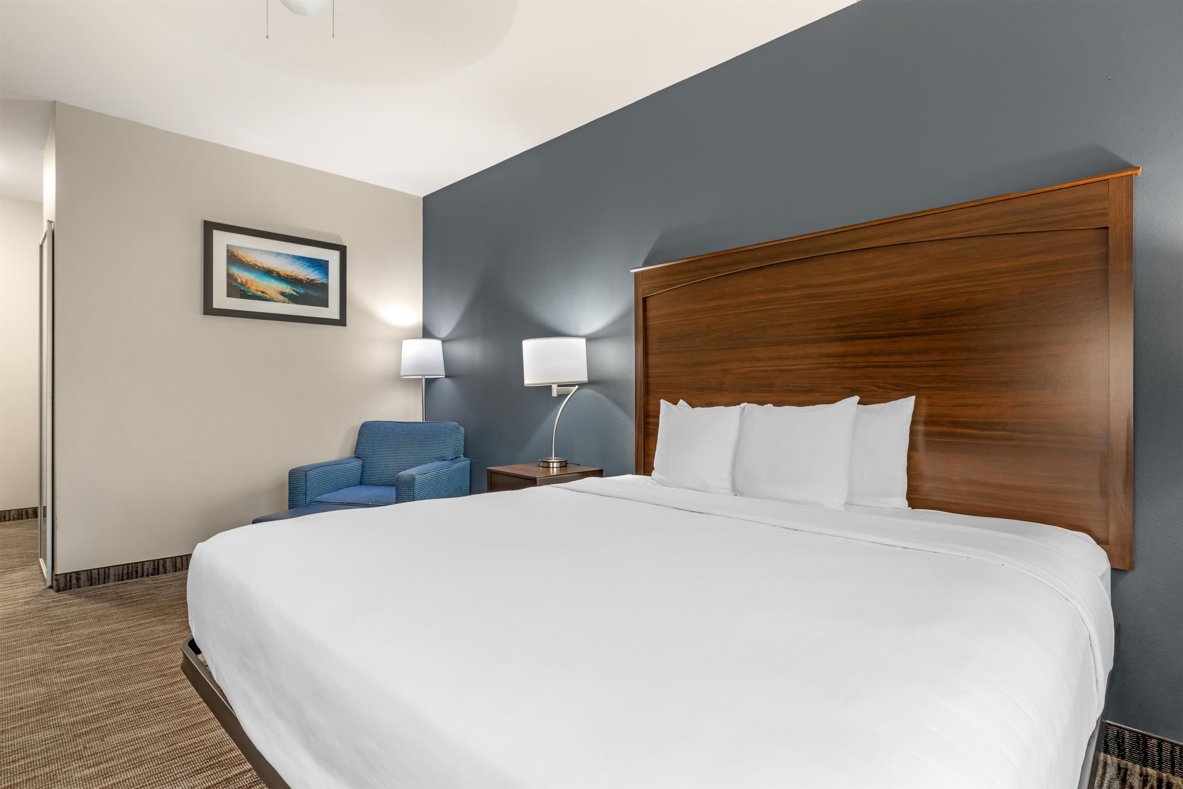 A hotel room with a large bed, a blue chair, a nightstand with a lamp, and a framed picture on the wall.