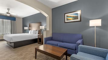 Habitación de hotel con cama, sofá, una mesita de centro, escritorio con lámparas, un cuadro en la pared y una lámpara de pie.