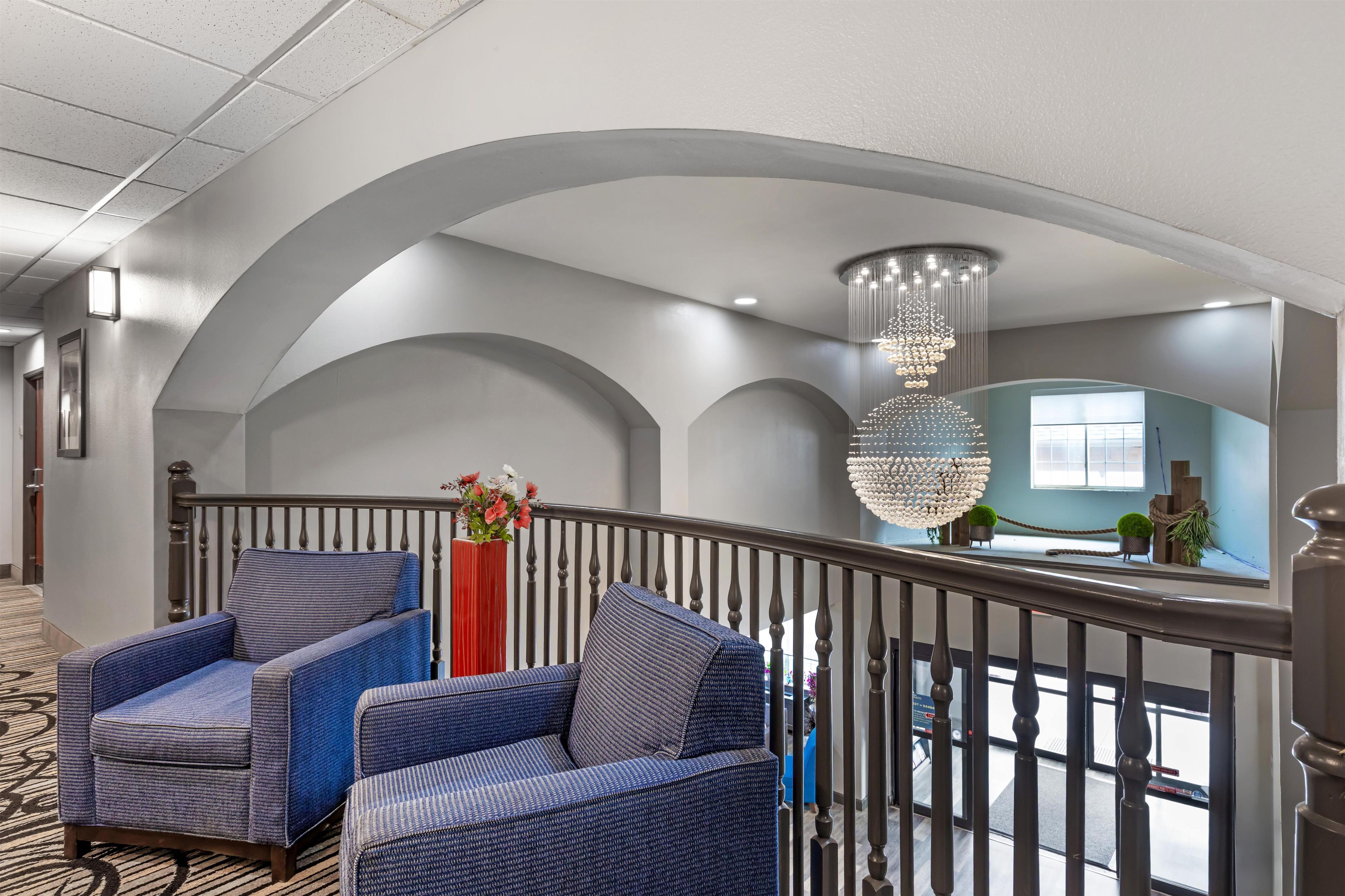 Um hall de hotel com design moderno, apresentando uma área de estar com poltronas estofadas em azul, um vaso vermelho com flores e um grande lustre.