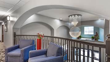 Um hall de hotel com design moderno, apresentando uma área de estar com poltronas estofadas em azul, um vaso vermelho com flores e um grande lustre.