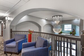 A hotel lobby with a modern design, featuring a seating area with blue upholstered chairs, a red vase with flowers, and a large chandelier.