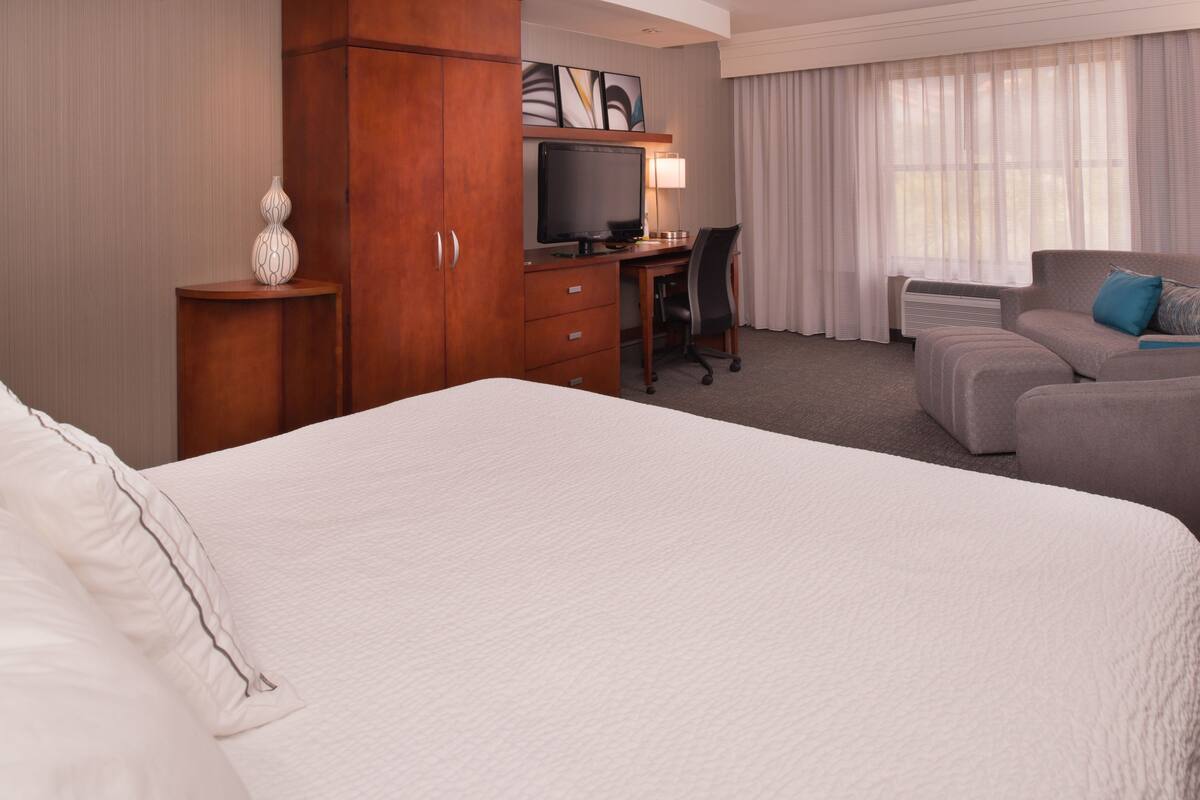A hotel room with a large bed, a desk with a computer, a chair, a sofa, and a TV.