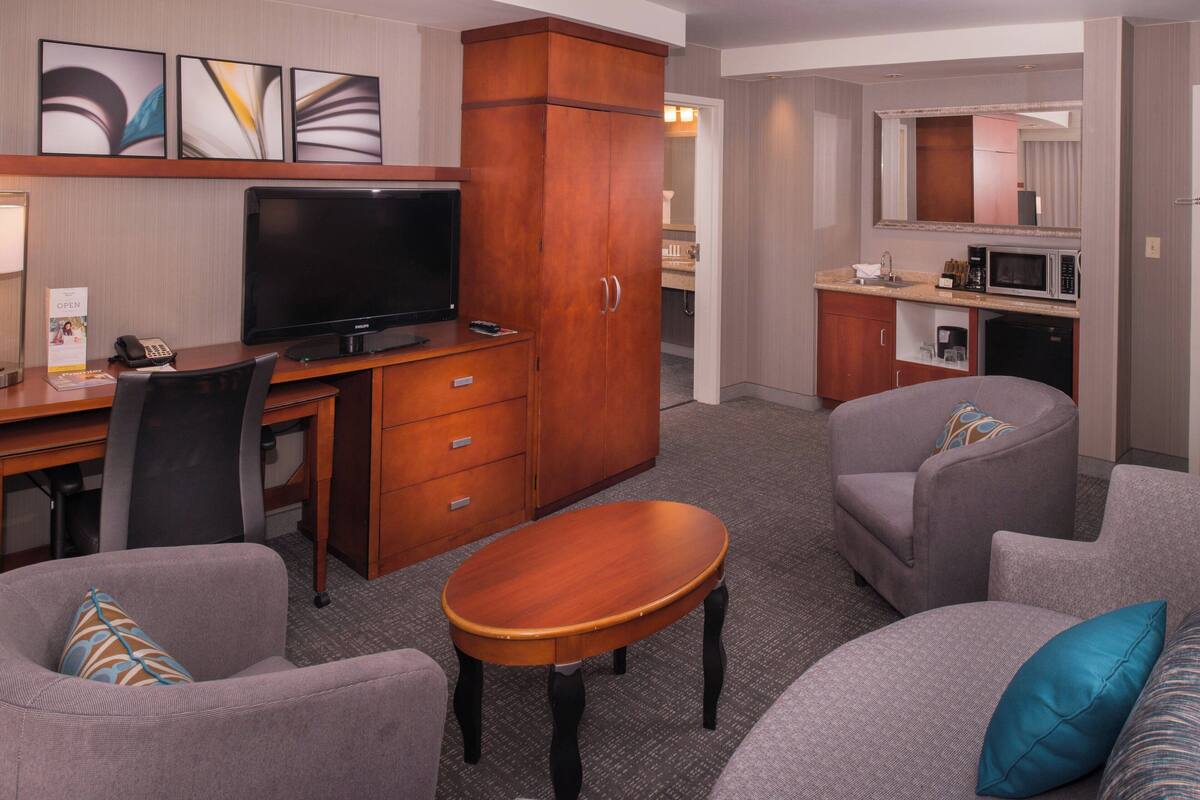 A hotel room with a television, a desk, a kitchenette, and two armchairs.