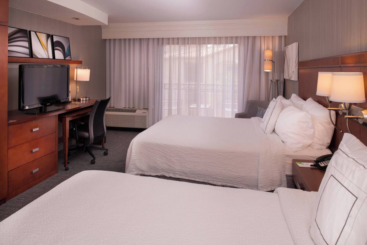 A hotel room with two beds, a desk with a television, a chair, a desk lamp, and a bedside table with a lamp.
