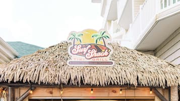 Um restaurante com temática tiki, com telhado de palha e um letreiro que diz 'Surf Shack TIKI GRILL'.