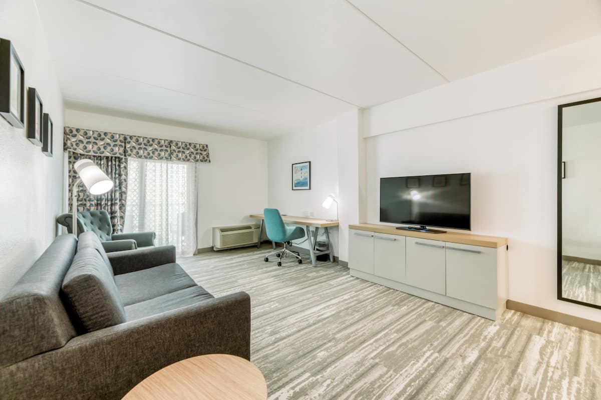 Quarto de hotel moderno com sofá, escrivaninha com cadeira, televisão e uma janela grande com cortinas.