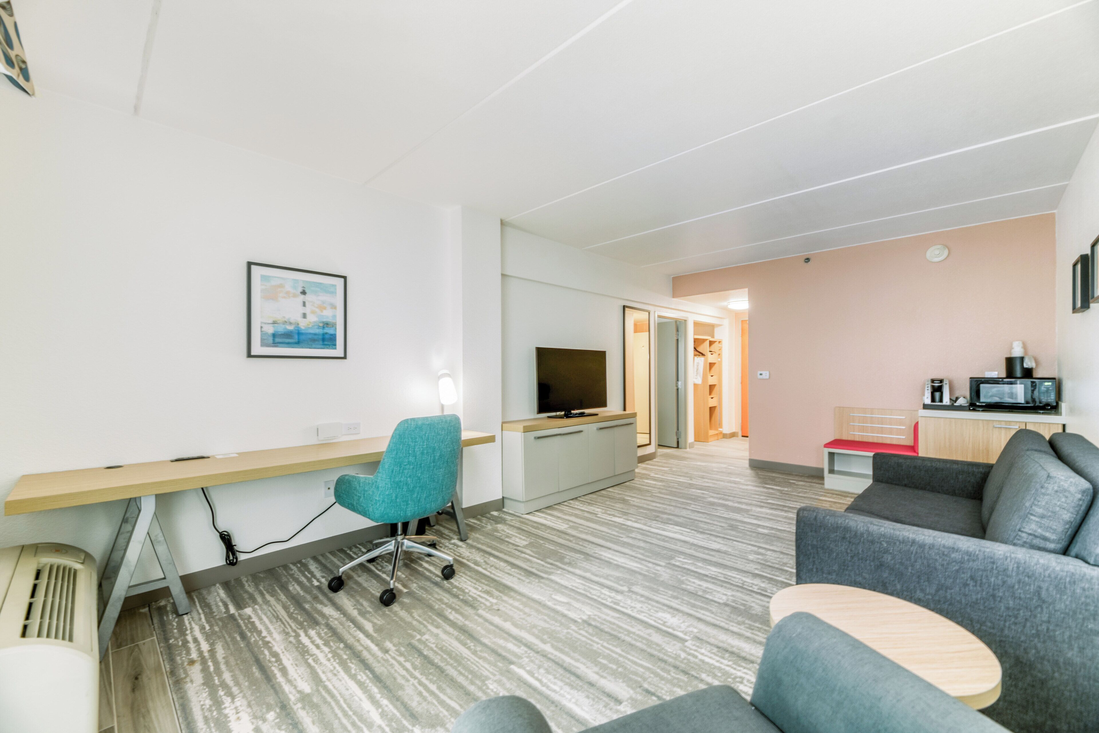 A modern hotel room with a desk, a television, a sofa, and a kitchenette.