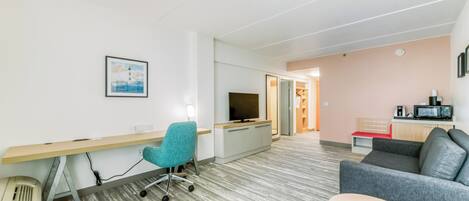 A modern hotel room with a desk, a television, a sofa, and a kitchenette.