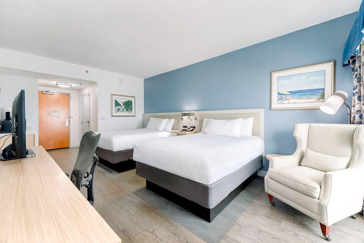 Ein Hotelzimmer mit zwei Betten, einem Schreibtisch, einem Sessel, einem Fernseher und einem Bild an der Wand.