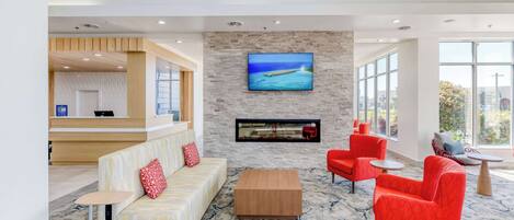 A modern lobby with a reception desk, a seating area with sofas and armchairs, a flat-screen TV mounted on a stone wall, and large windows offering views of the outdoors.