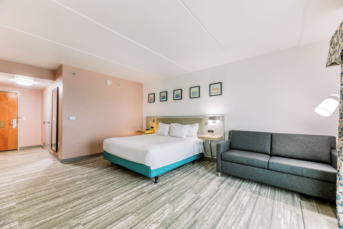 Quarto de hotel com uma cama, um sofá cinza, uma mesa de cabeceira e uma luminária fixada na parede.