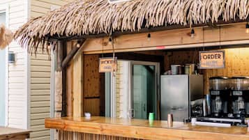 Um bar tiki com telhado de palha, uma placa com o dizer 'Sully Crack Tiki Grill' e um balcão com a indicação 'FAÇA O PEDIDO AQUI'.