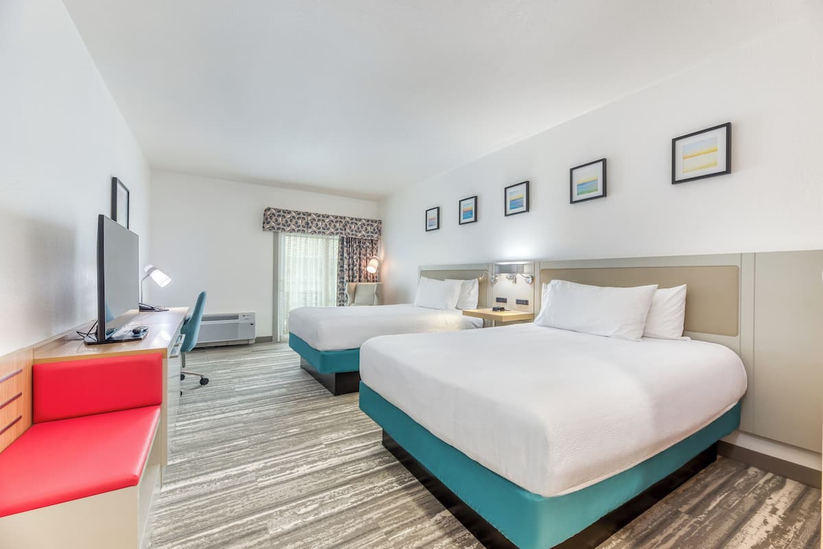 Quarto de hotel com duas camas, uma escrivaninha com computador, um banco vermelho e uma janela com cortinas.