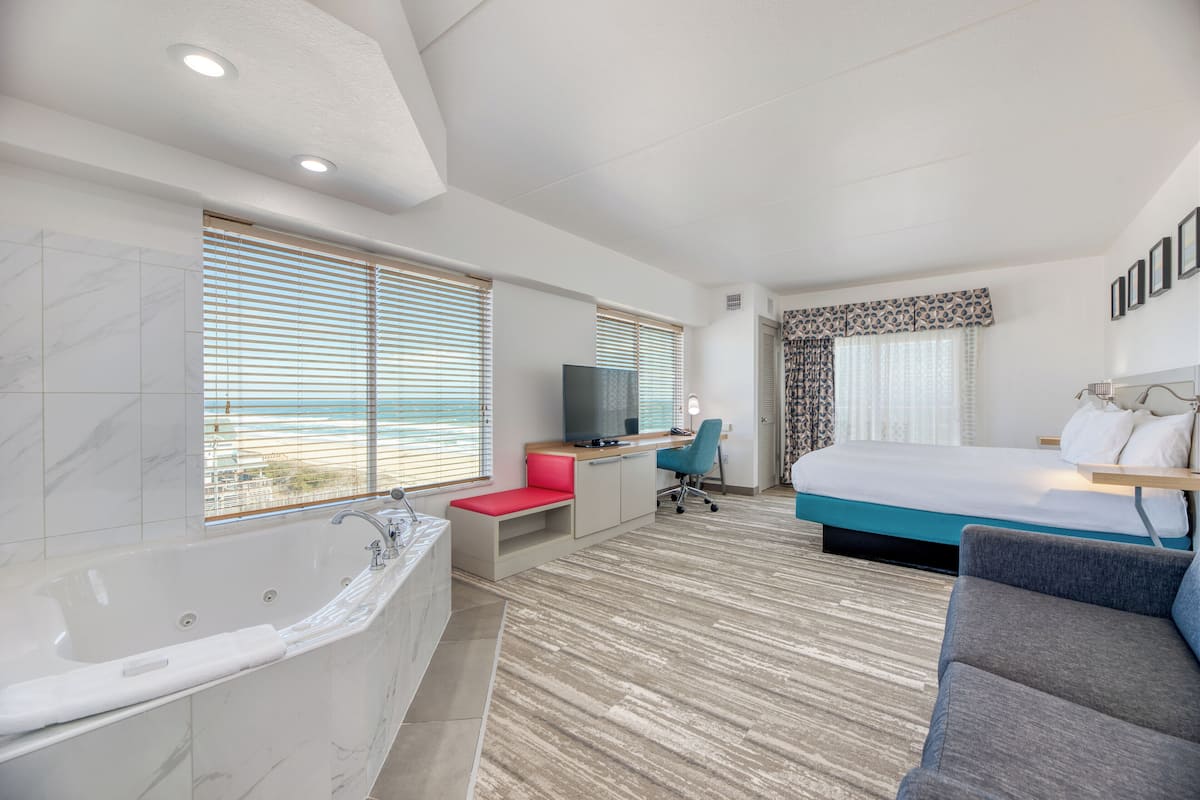Quarto de hotel moderno com uma cama grande, uma escrivaninha, uma cadeira, uma banheira e janelas amplas com cortinas.