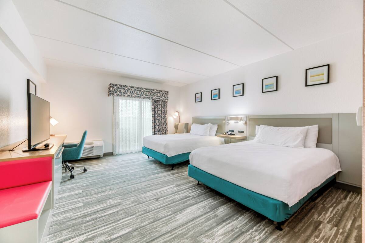 Quarto de hotel com duas camas, uma escrivaninha com cadeira, televisão e janela com cortinas.
