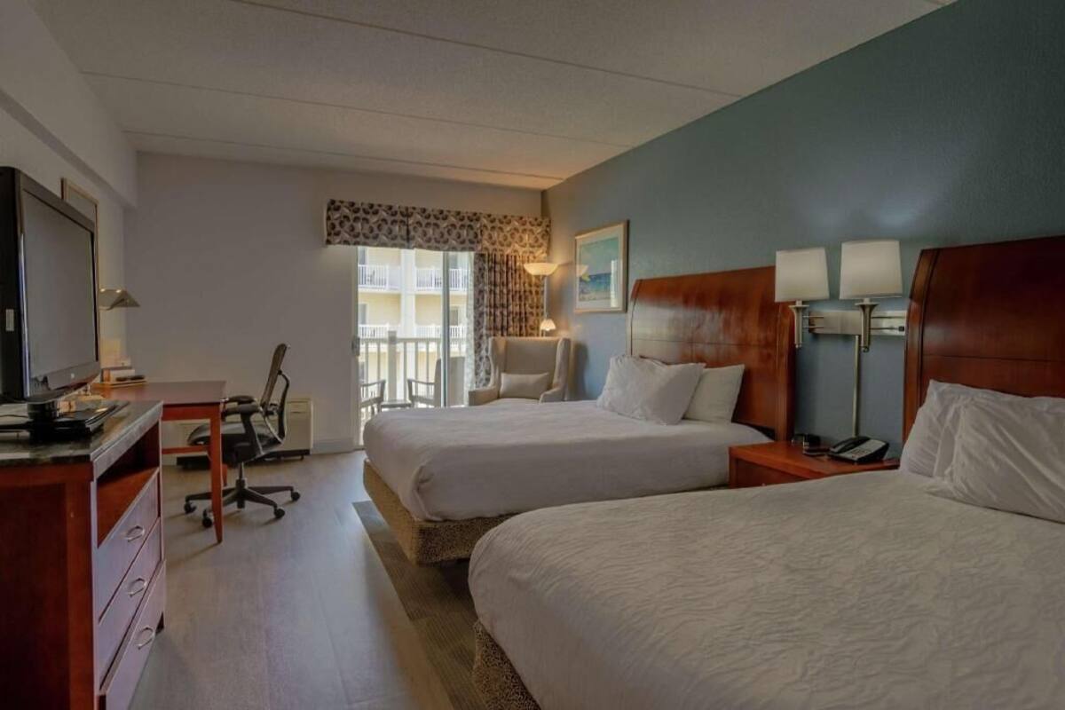 Quarto de hotel com duas camas, uma escrivaninha com computador, uma cadeira e vista para uma varanda.