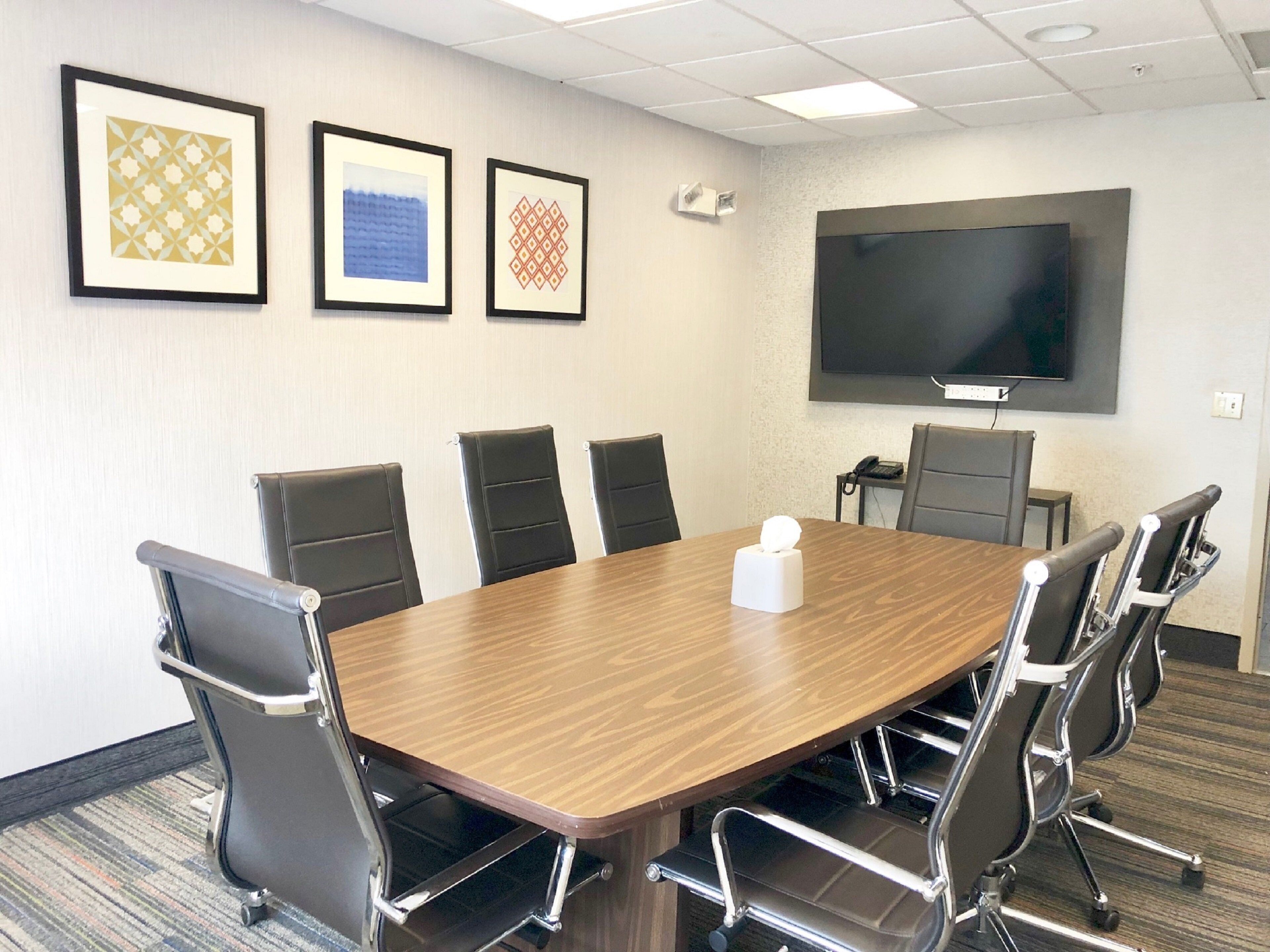 A conference room with a large table, six chairs, a television on the wall, and three framed artworks.