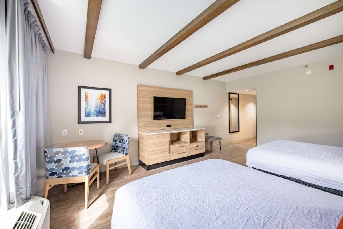 A hotel room with a bed, a TV, a small table with chairs, and a window with curtains.