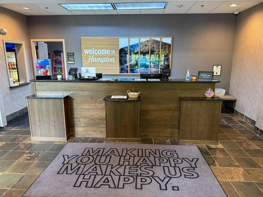 Zona de recepción de un hotel con un cartel de 'Welcome to Hampton', un mostrador de recepción, un cartel de 'Stay Only', un cartel de 'Manager On Duty', un cartel de 'Hilton Honors' y una alfombra con la inscripción 'Making You Happy'.