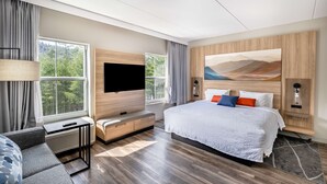 A modern hotel room with a large bed, a flat-screen TV, a seating area with a table and bench, and a window offering a view of trees. - Hampton Inn & Suites Cashiers Sapphire Valley (Sapphire)