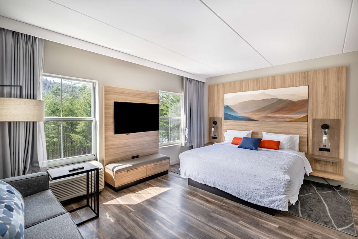 A modern hotel room with a large bed, a flat-screen TV, a seating area with a table and bench, and a window offering a view of trees.