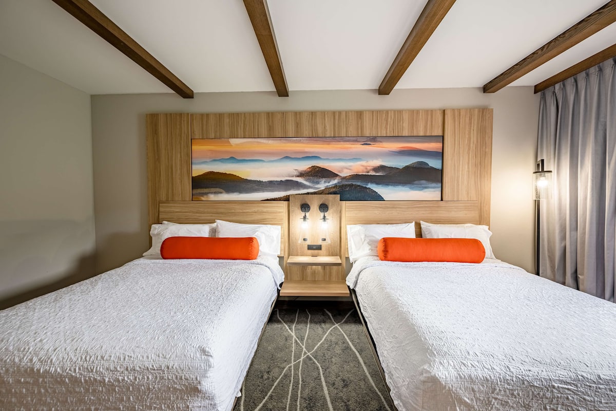 A hotel room with two beds, a large painting on the headboard, and a nightstand with lights.