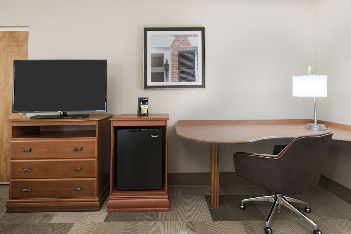 A hotel room with a flat-screen TV, a mini-fridge, a desk with a chair, and a framed picture on the wall.