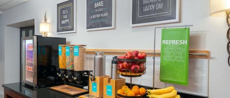 Un bar où l’on peut prendre le petit-déjeuner, avec une machine à café, des corbeilles de fruits et un panneau vert.