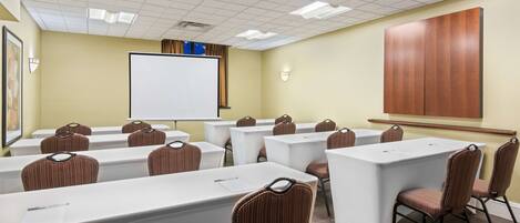 A conference room with white tables and chairs, a projector screen, and a large wooden panel on the wall.