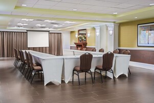 Un salón de conferencias con una larga mesa, sillas, una pantalla de proyector y un atril con un jarrón de flores.