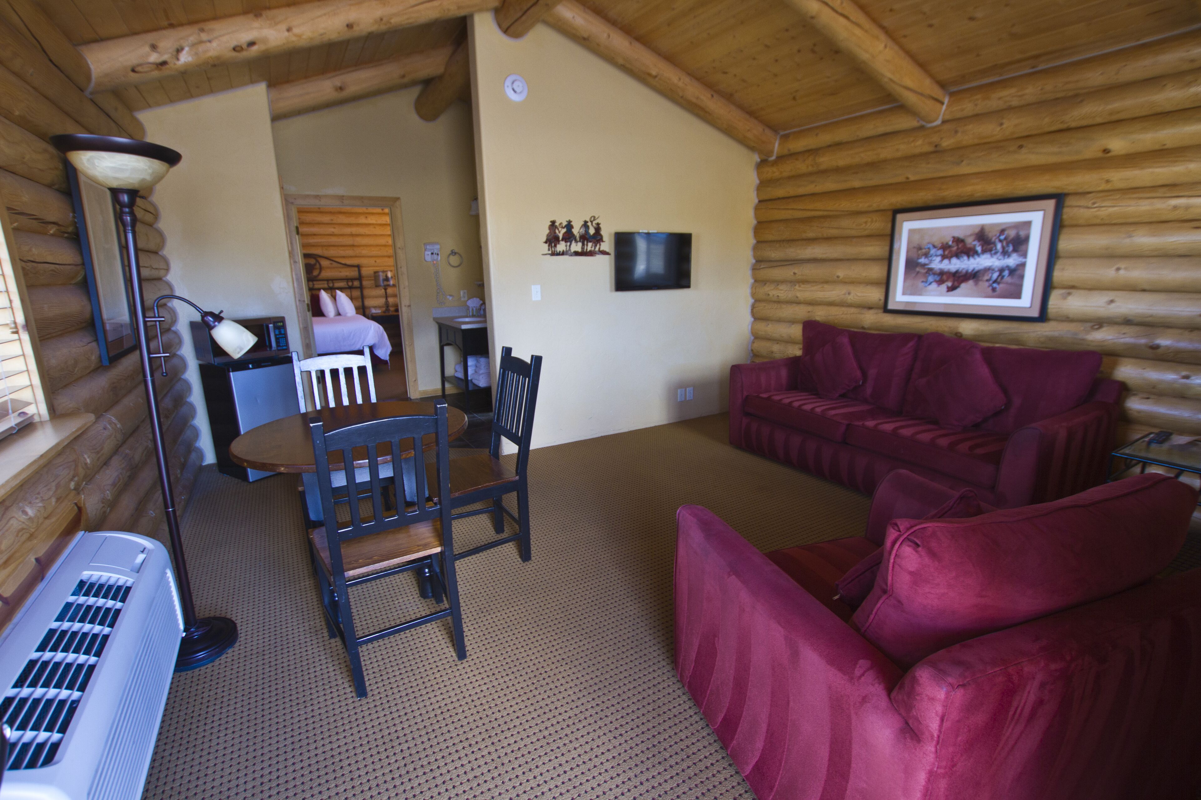 Interior de una cabaña de madera con piso de madera, un sofá rojo, una mesa de comedor de madera con sillas, una lámpara y un televisor en la pared.