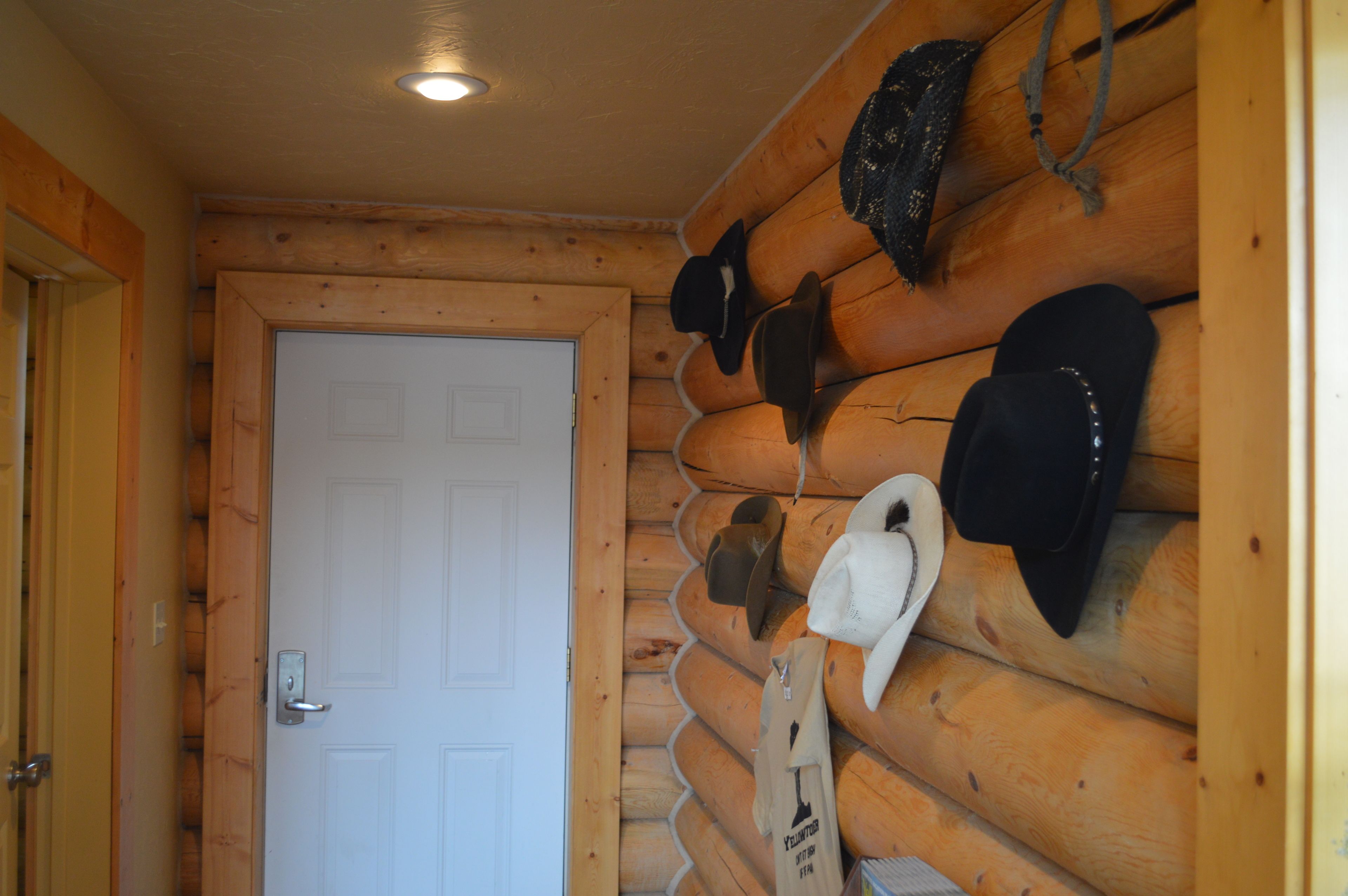 L’intérieur d’une cabane en rondins, avec une porte blanche, des murs en bois, et plusieurs chapeaux, dont un chapeau de cow-boy, accrochés au mur.