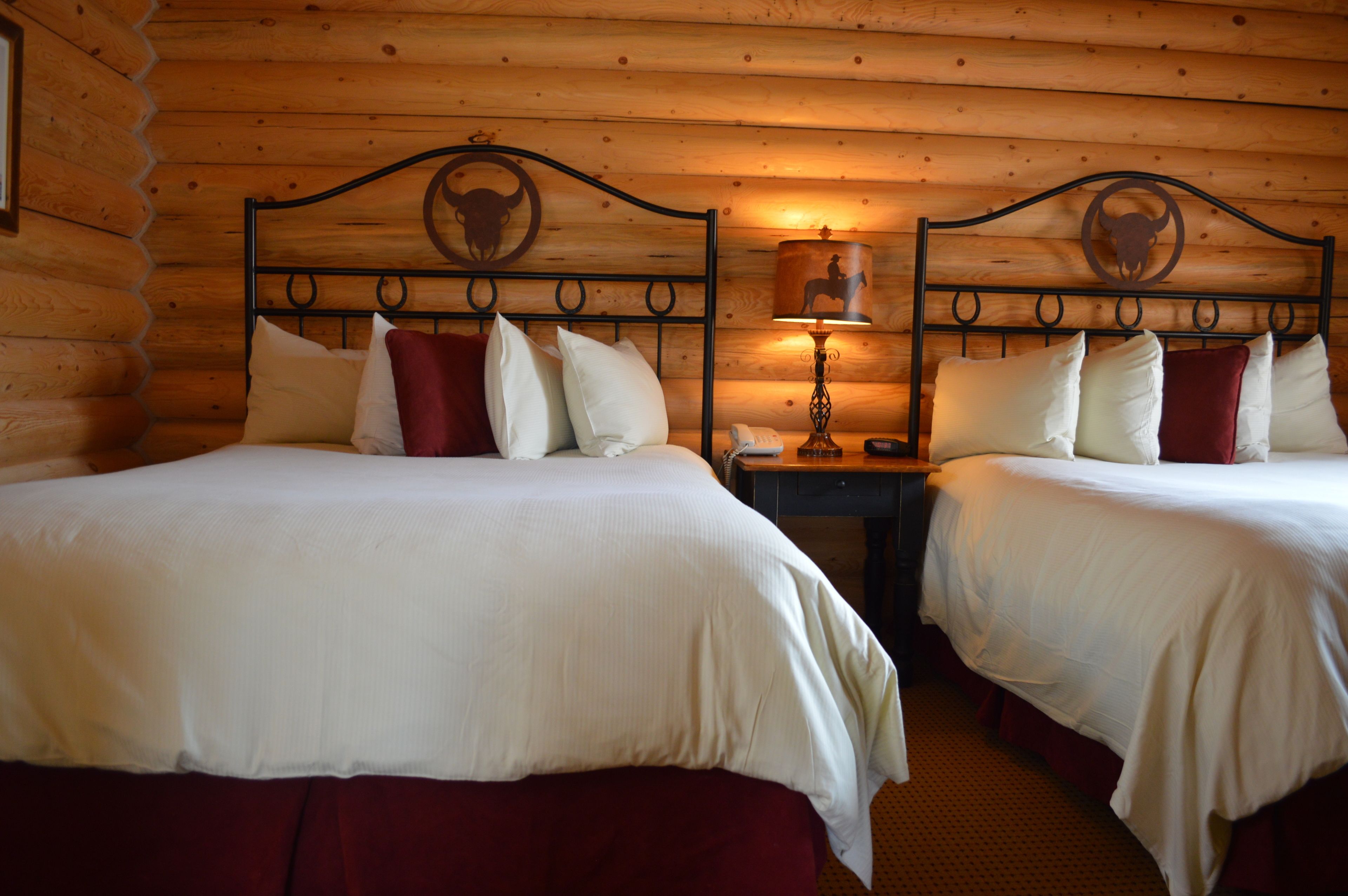 Een kamer in chaletstijl met twee bedden, elk voorzien van wit beddengoed en rode kussens, een houten hoofdbord met een ontwerp van een stierenhoofd, een nachtkastje met een lamp en een telefoon.