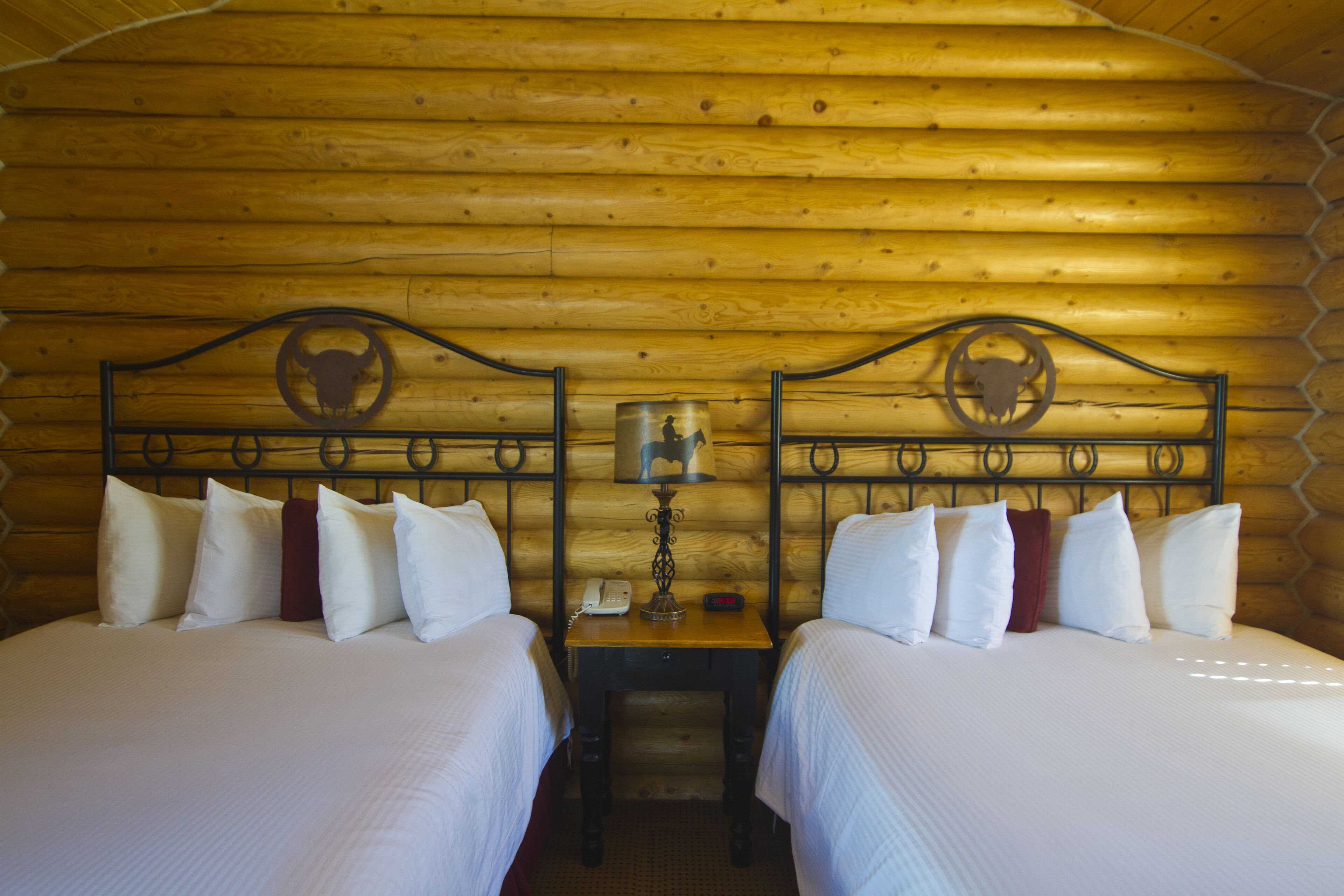 A wooden cabin room with two beds, each with white bedding and a bull head design on the headboard. There is a nightstand with a lamp, a phone, and a red object.