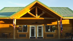 A log cabin with a green metal roof, two doors with signage, and American flags. - Cody Cowboy Village (Cody)