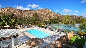 A swimming pool with lounge chairs, a tennis court, and mountains in the background.