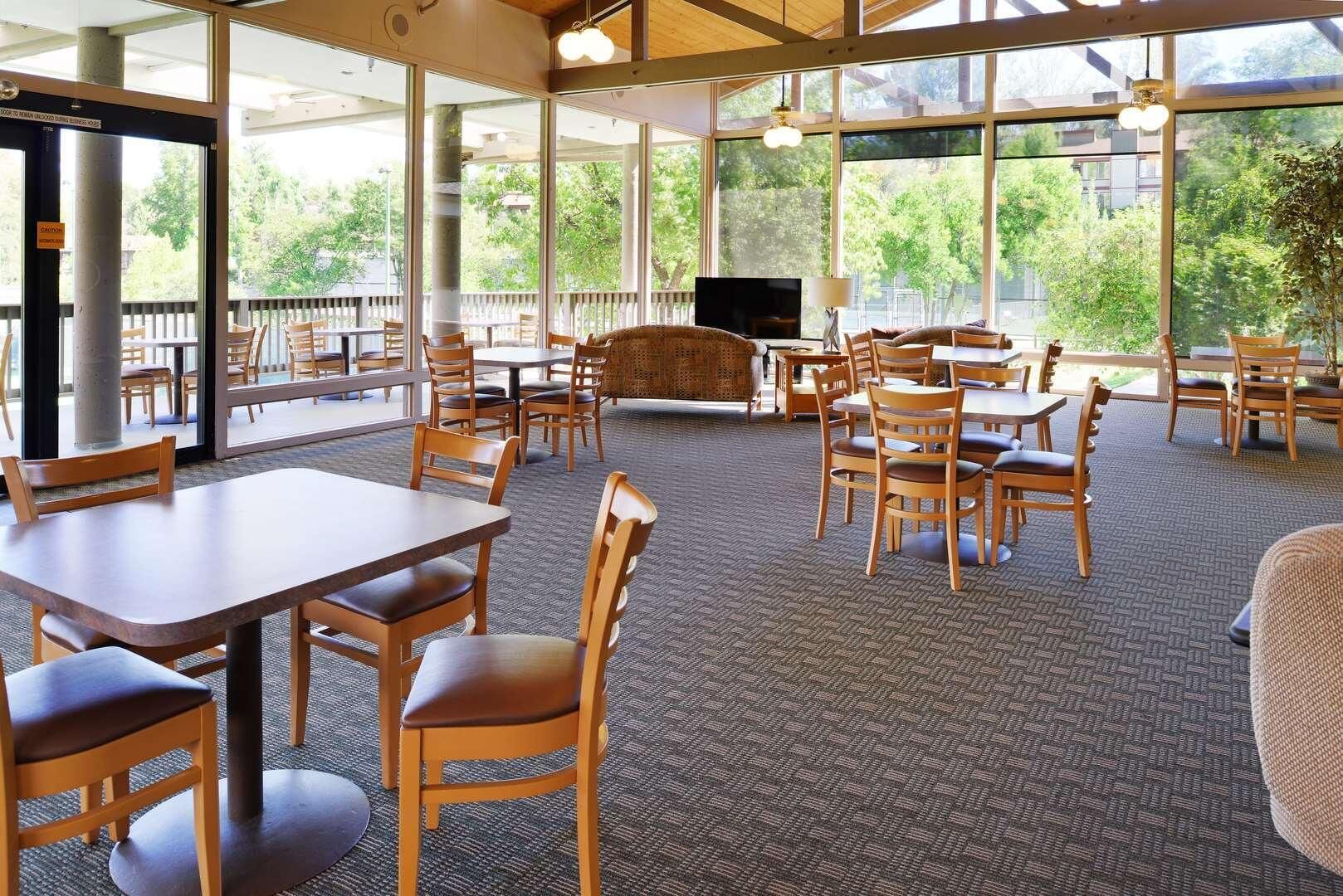 A spacious indoor area with multiple tables and chairs, large windows offering a view of greenery outside, and a television on a stand.