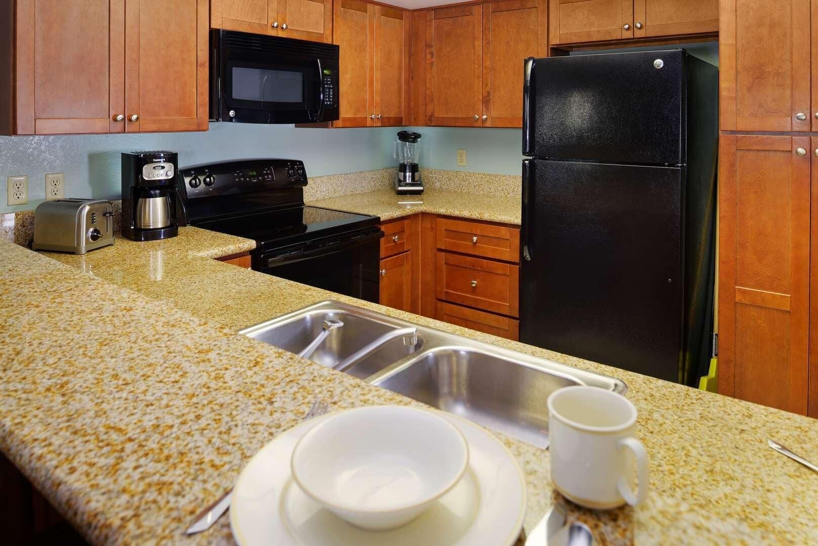 Una cocina con armarios de madera, un microondas negro, un refrigerador negro, una cafetera y un lavamanos.