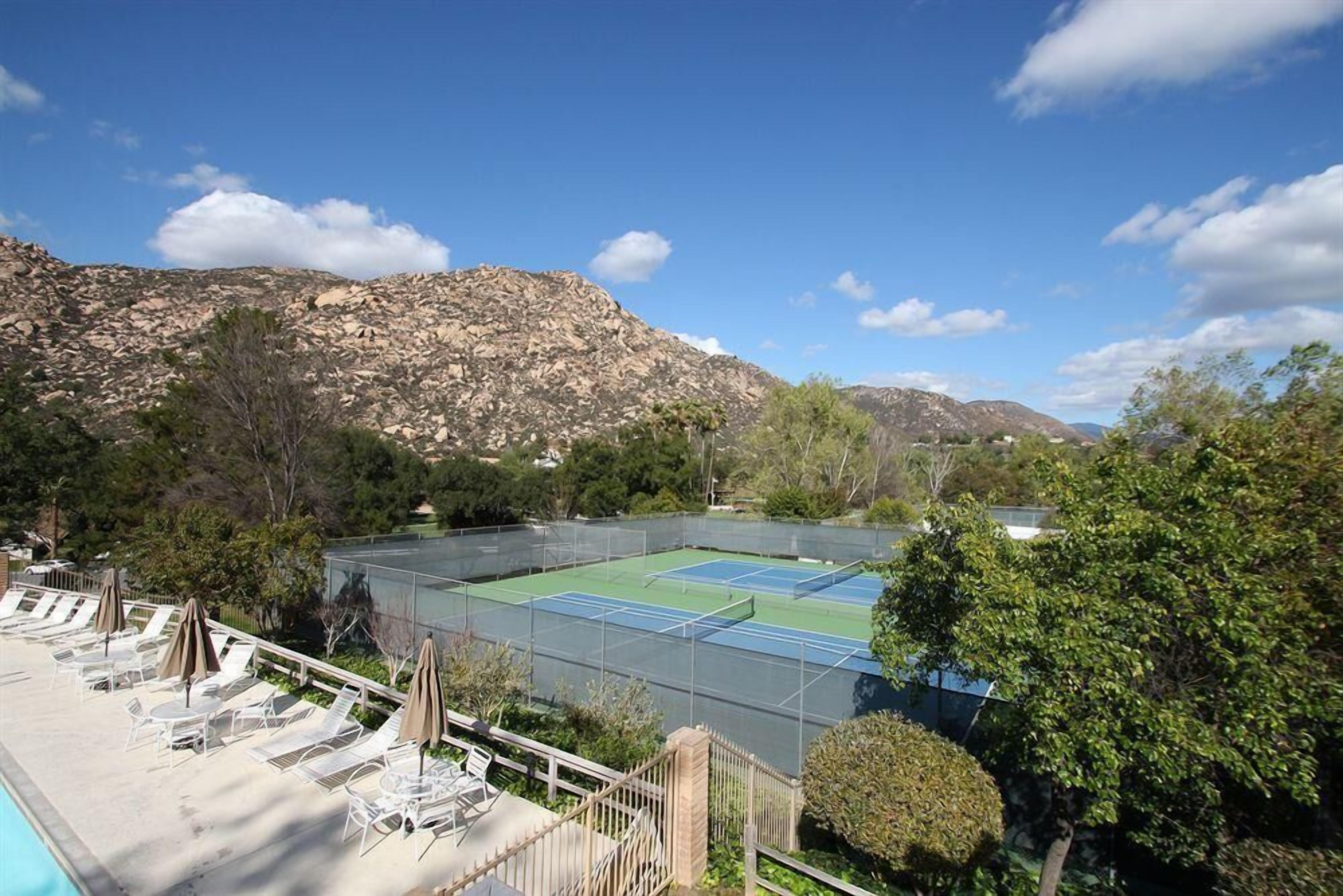 Un court de tennis avec des chaises longues et une piscine, le tout entouré de montagnes et d’arbres.