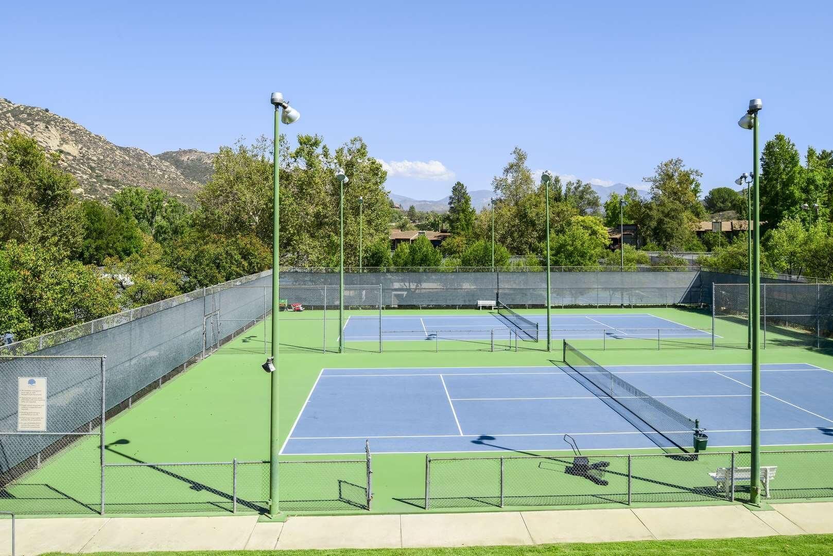 A tennis court with multiple courts, surrounded by a fence, and with trees and mountains in the background.