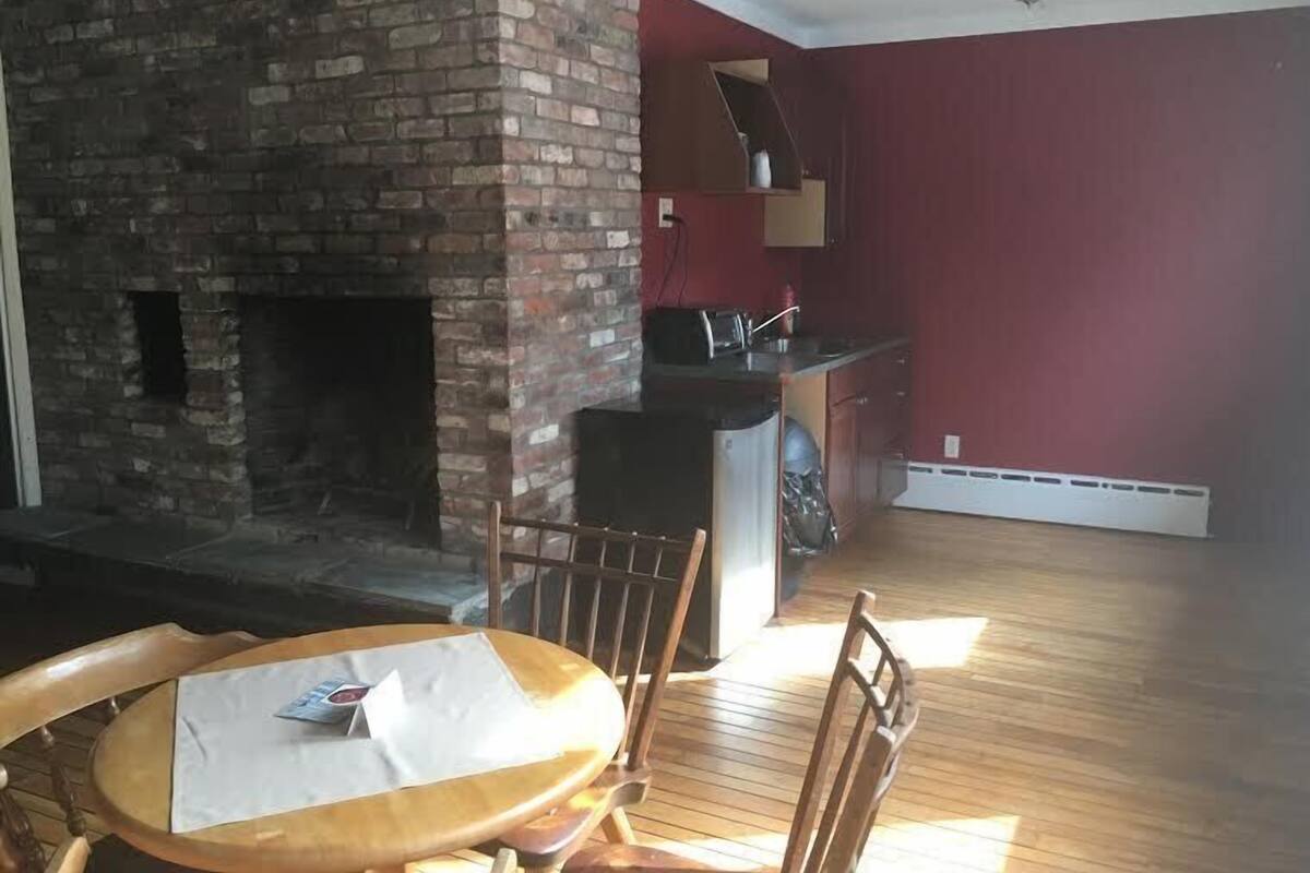 A room with a brick fireplace, a dining table with chairs, and a kitchen area with red walls and wooden flooring.