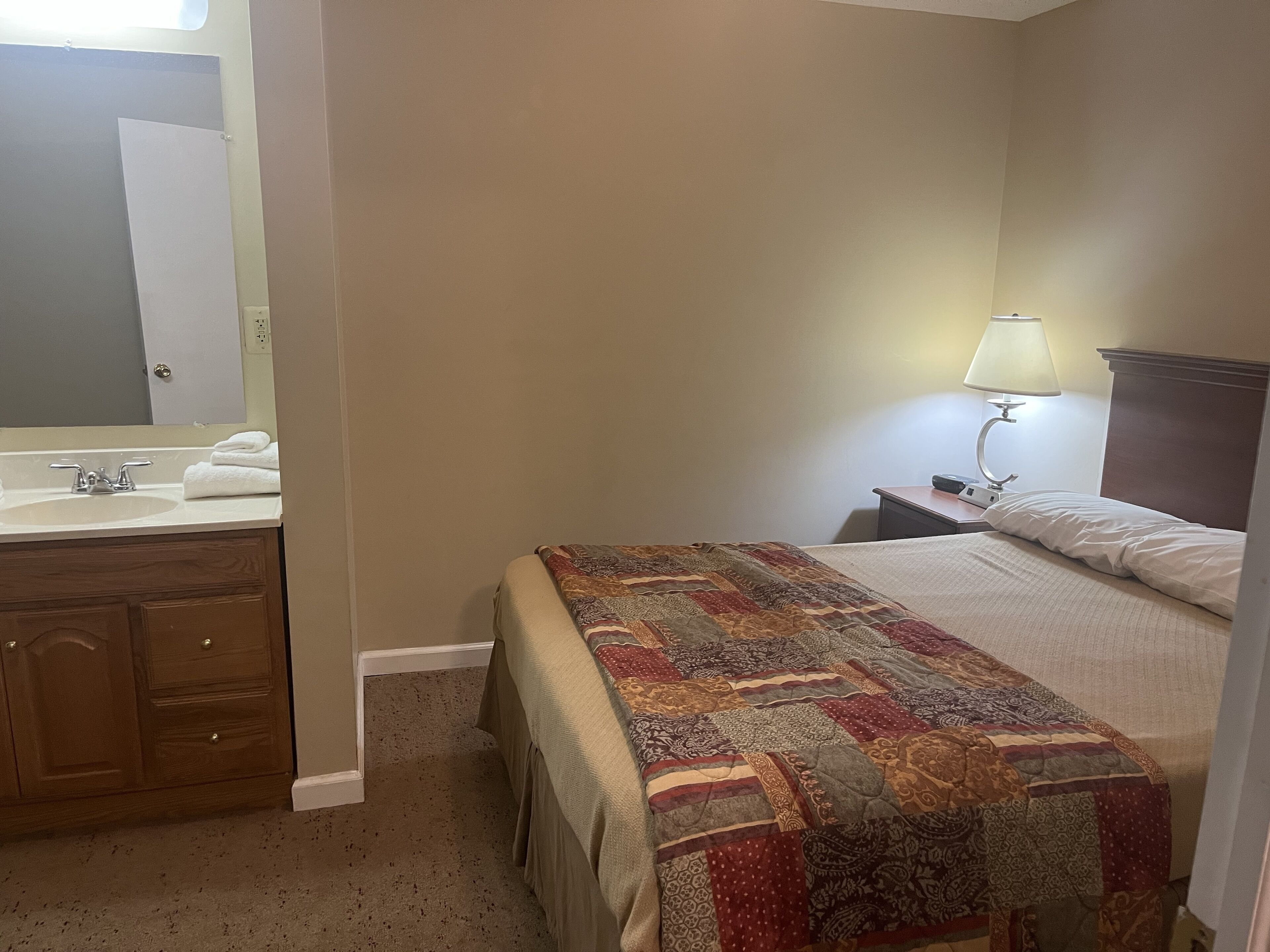 Habitación de hotel con cama, mesita de noche con lámpara, baño con lavamanos y espejo, y una puerta.
