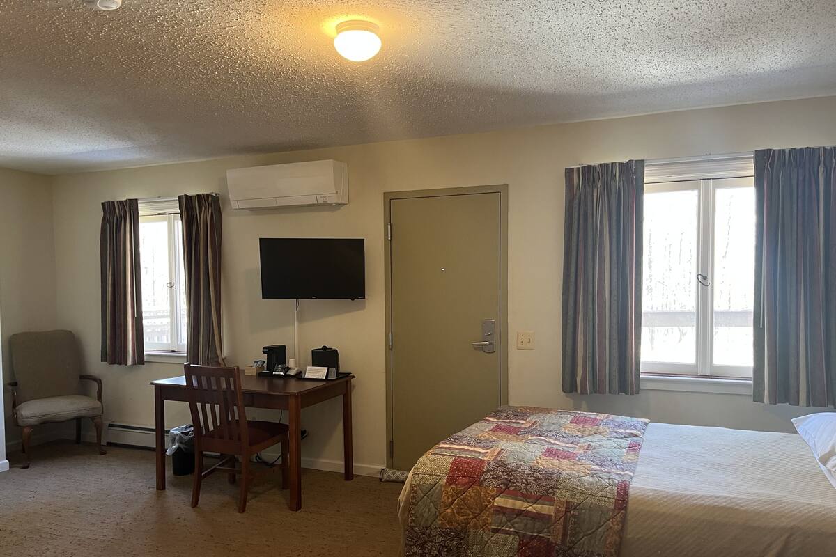 A hotel room with a bed, a desk with a chair, a television, and an air conditioning unit.