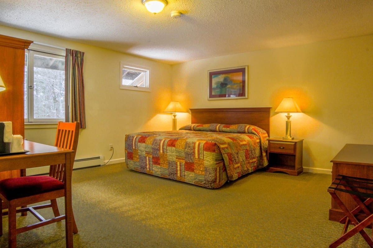 A hotel room with a bed, two bedside lamps, a desk with a chair, a window with curtains, and a painting on the wall.