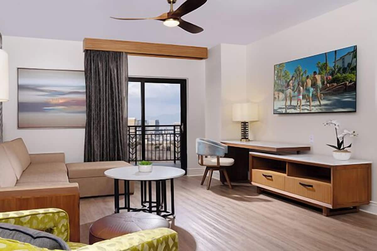 A modern hotel room with a sofa, armchair, dining table, and a balcony with a city view.