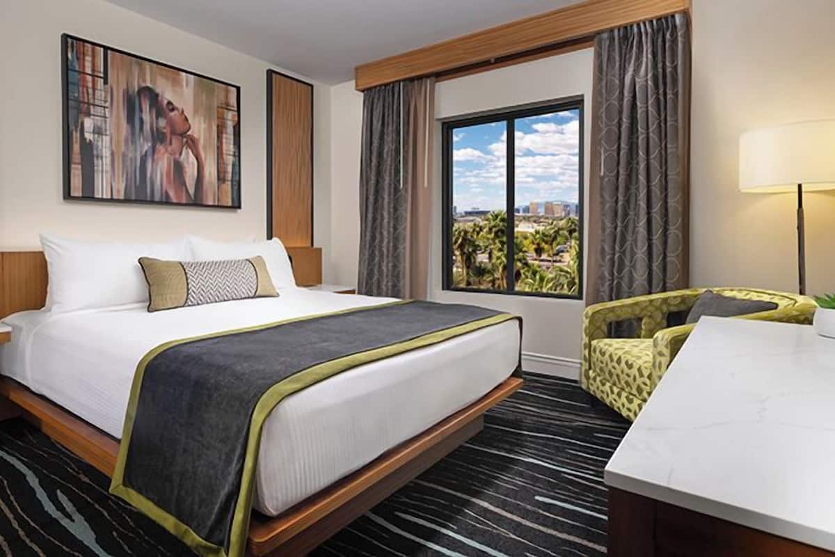 A hotel room with a large bed, a desk, a chair, a window with a city view, and a painting on the wall.