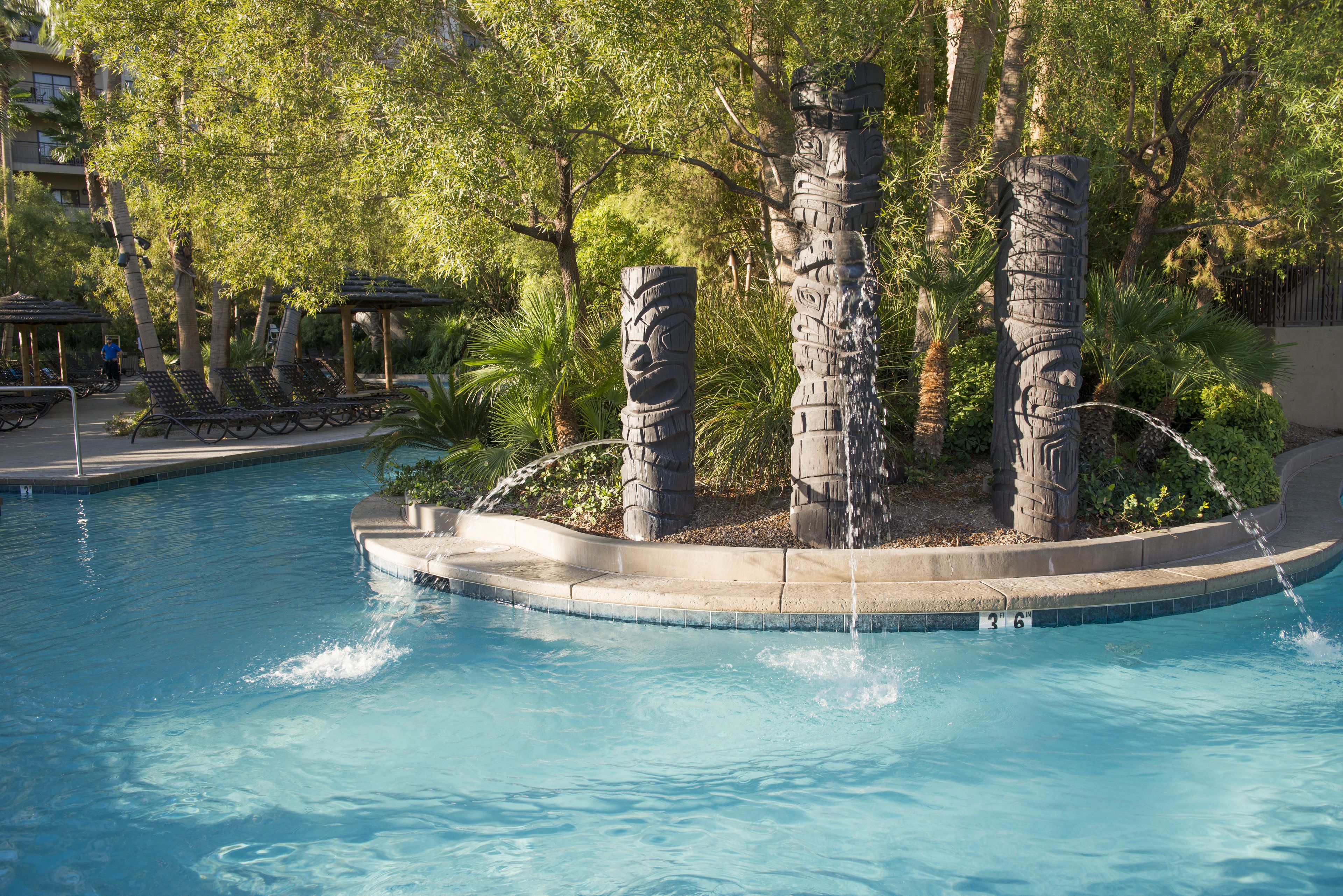 A swimming pool with a stone sculpture in the center, surrounded by palm trees and lounge chairs.